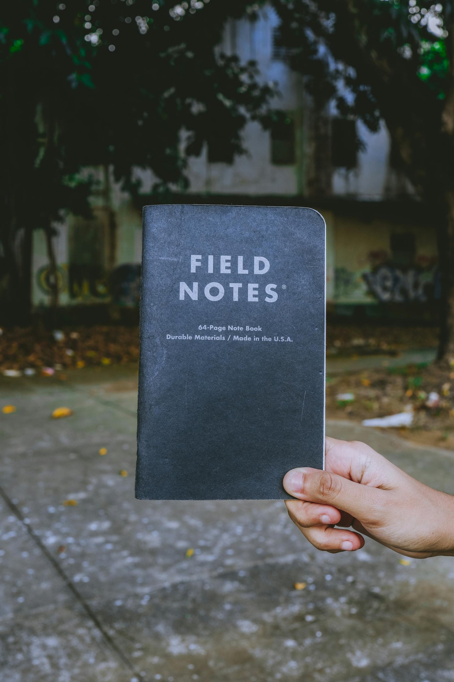 Picture of a note book titled 'Field Notes' being held by a person's hand with a dark tree and building behind the image. The image signifies Horticulture Consults in the field.